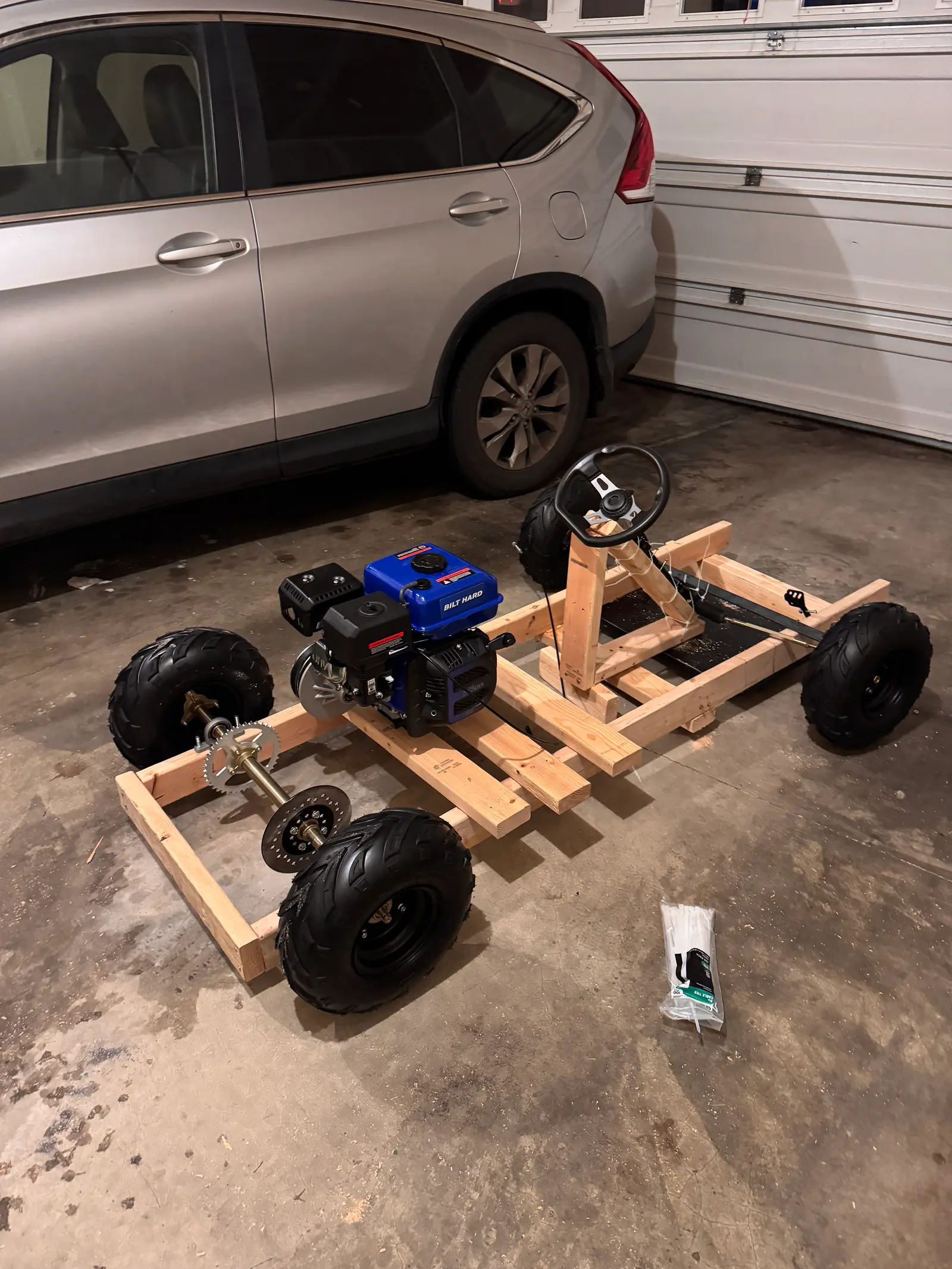 The finished wood go-kart in a garage: timber frame, blue Predator engine, steering wheel and four knobbly tyres